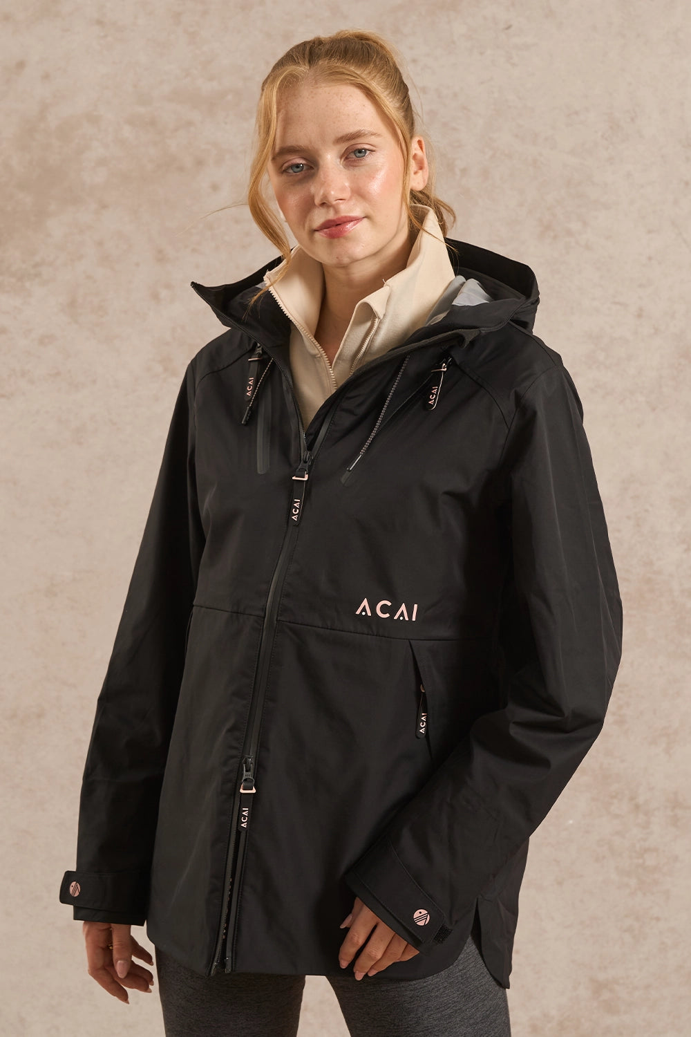 A woman wearing a black hooded jacket with a beige collar and 'ACAI' printed on the chest, posed against a textured beige background.
