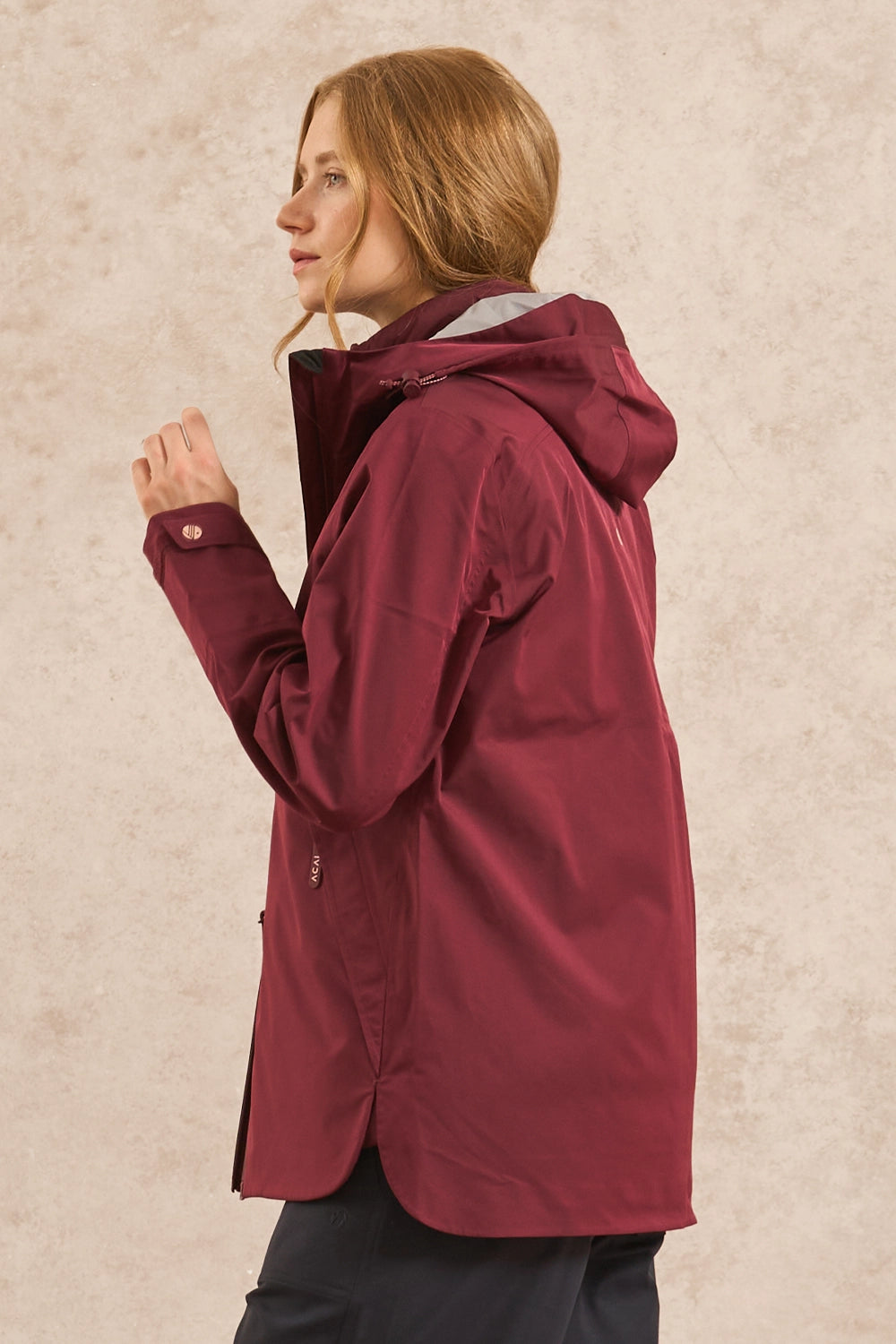 A woman wearing a maroon hooded rain jacket, shown in profile against a textured beige background.