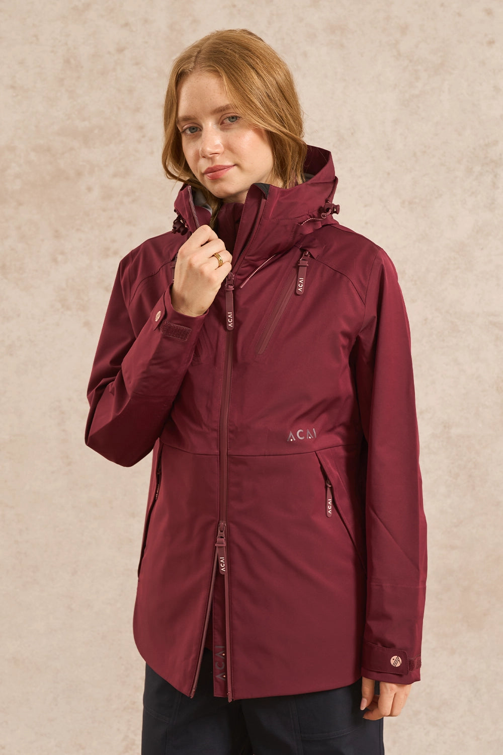 A woman wearing a maroon hooded waterproof jacket with a full front zipper and zippered hand pockets, standing against a textured beige background.