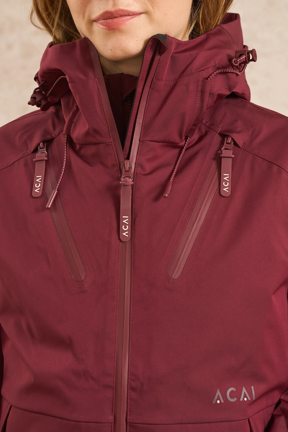 A close-up of a person wearing a maroon hooded technical jacket with multiple zippered pockets and visible AAI branding on the chest and zipper pulls.