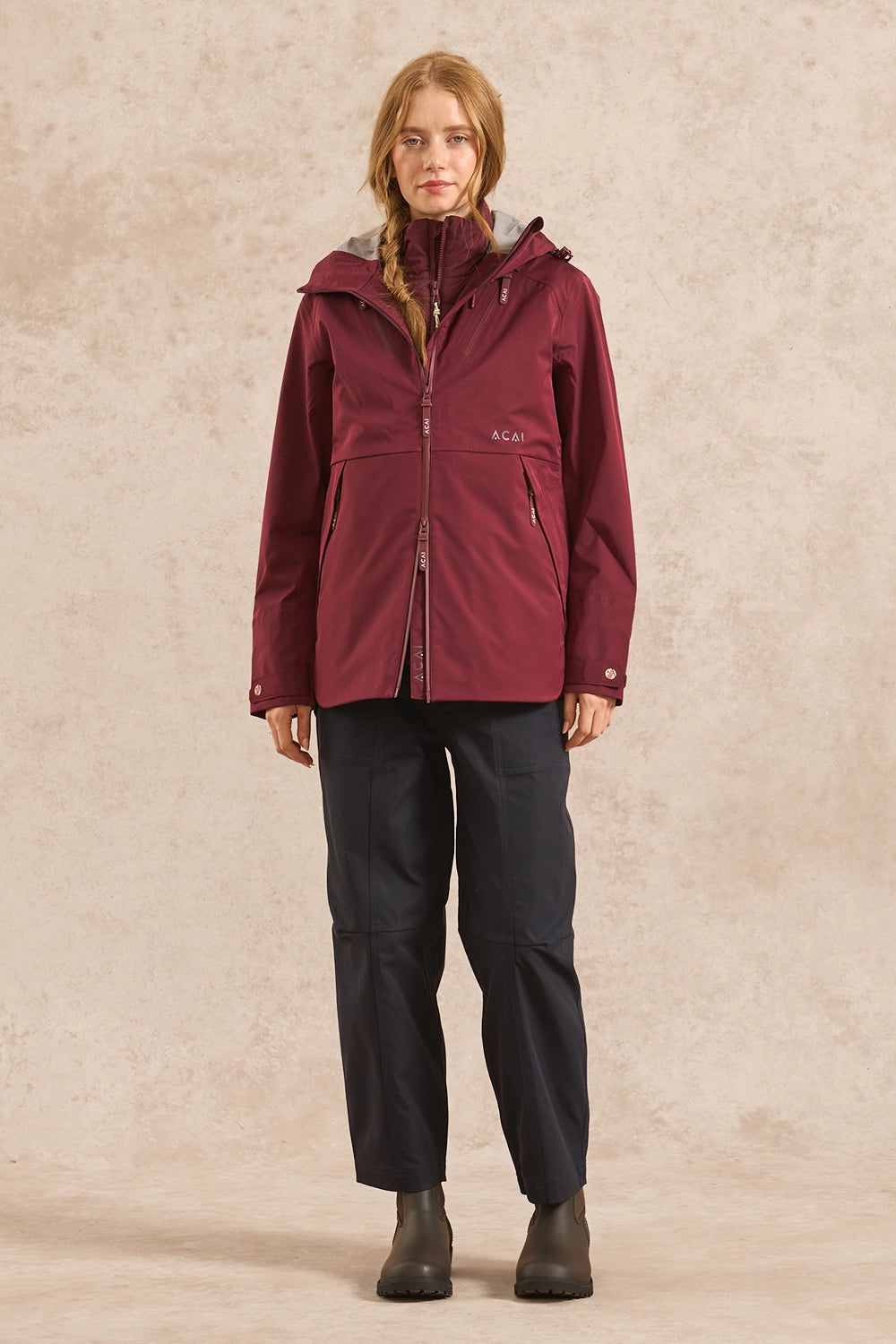 A woman modeling a maroon hooded technical jacket with a gray inner hood lining, paired with dark pants and boots, standing against a textured beige studio backdrop.