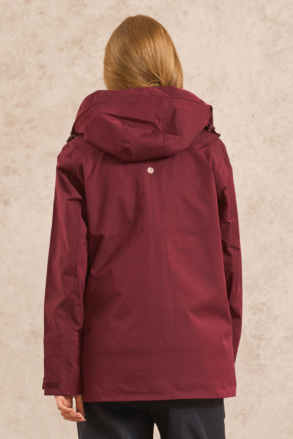 A woman viewed from behind wearing a hooded jacket in deep burgundy, set against a textured beige background.