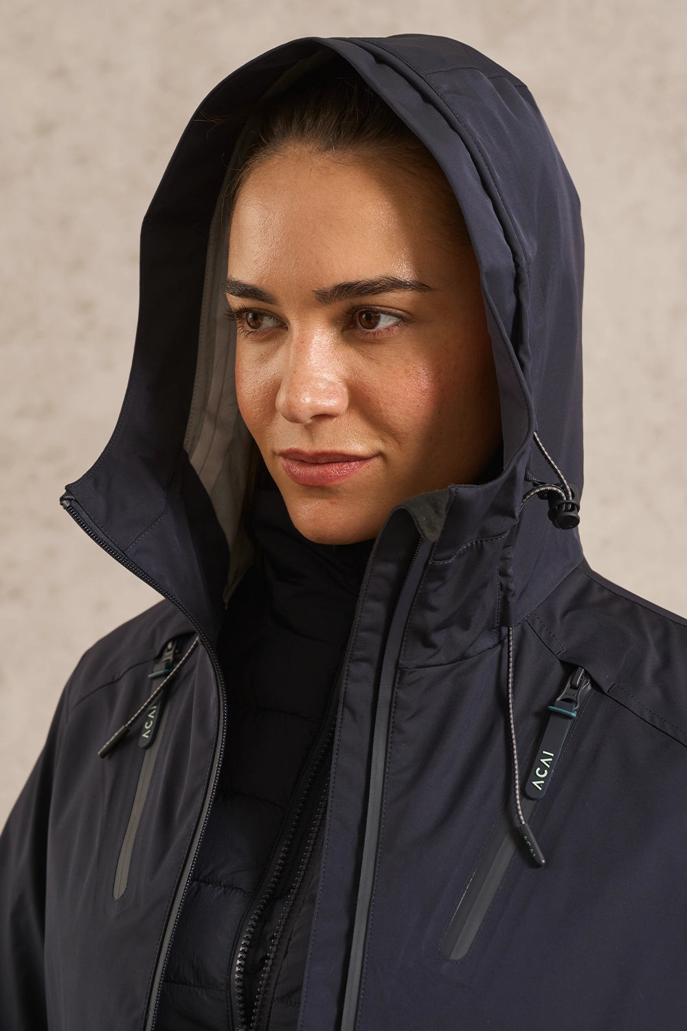 A close-up of a woman wearing a dark navy hooded jacket with a high collar and front zipper, photographed against a neutral textured beige background.