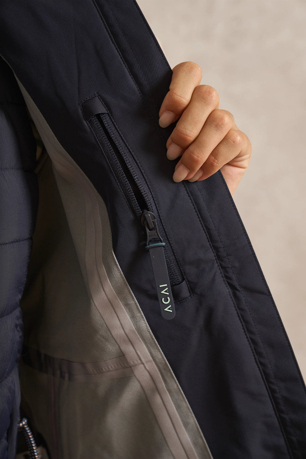 Close-up of a person's hand holding open a dark navy puffer jacket to reveal a gray inner layer and a zippered pocket with a visible ACAI.