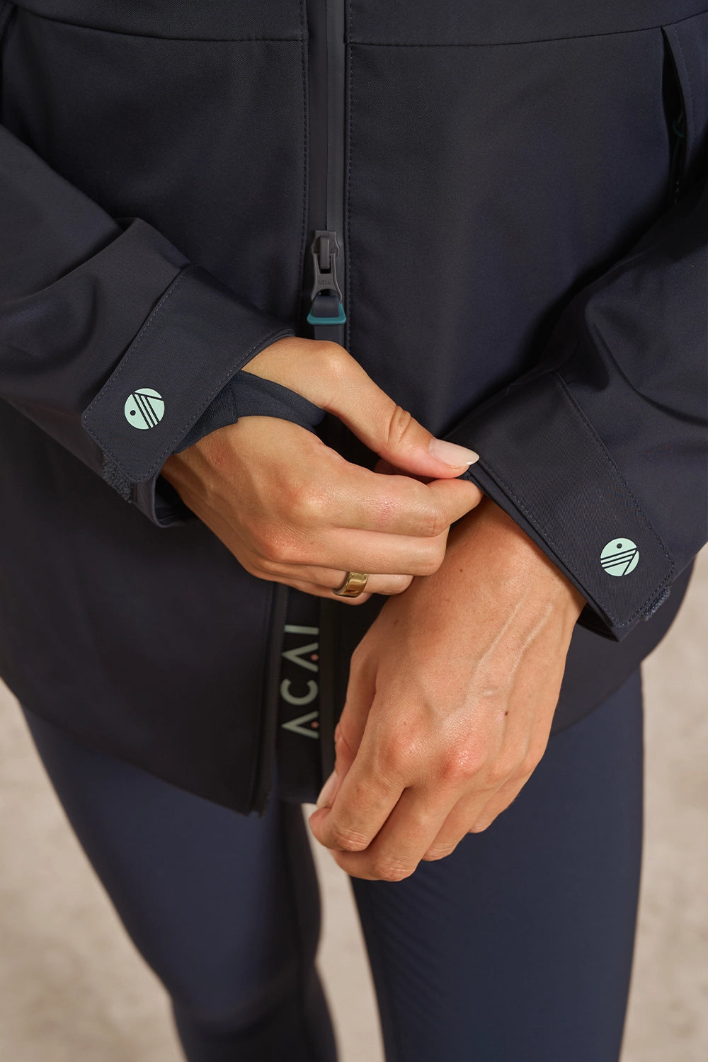 Close-up of a person wearing a dark navy athletic jacket and matching leggings, adjusting the cuff of the jacket. The jacket has a front zipper, long sleeves, and circular logo patches on the cuffs.