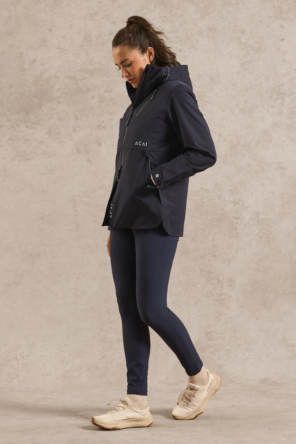 A woman wearing a navy blue hooded jacket with 'ACAI' printed on the chest, navy leggings, and beige sneakers, standing against a textured beige studio background.