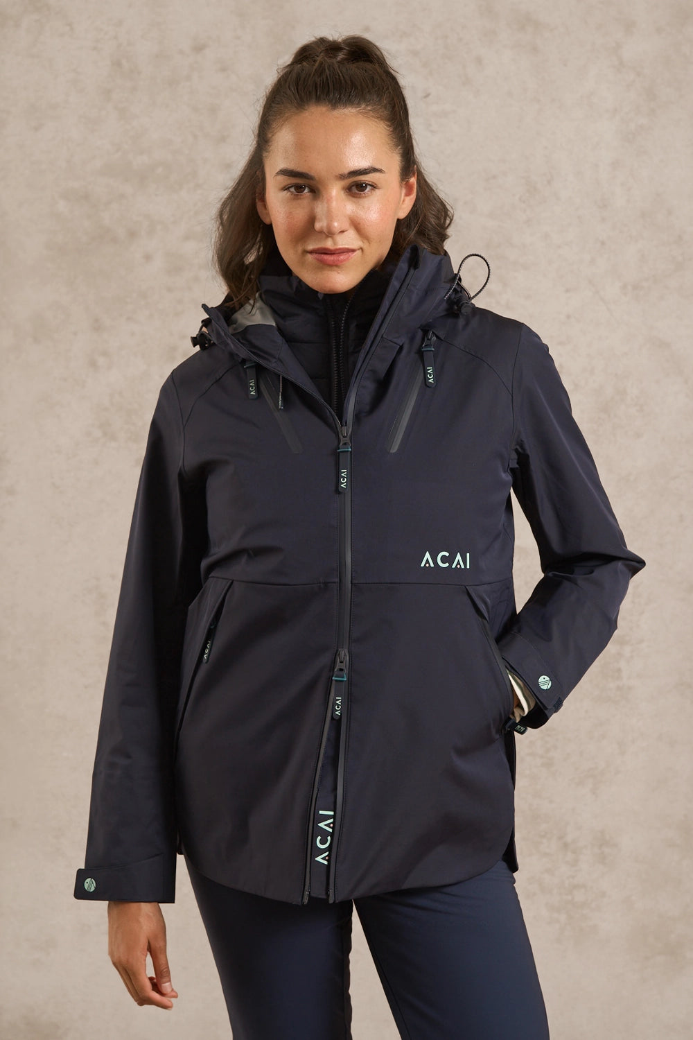 A woman modeling a dark navy Acai outdoor jacket with a high collar, front zipper, and zippered pockets, set against a textured beige studio background.