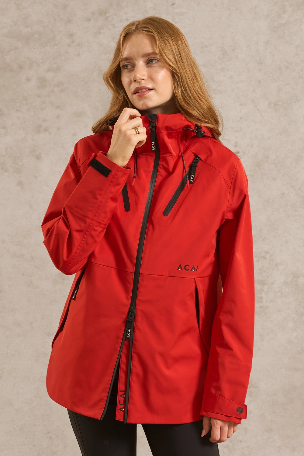 A woman wearing a bright red hooded jacket with black zippers and black logo details, standing against a textured light gray wall.