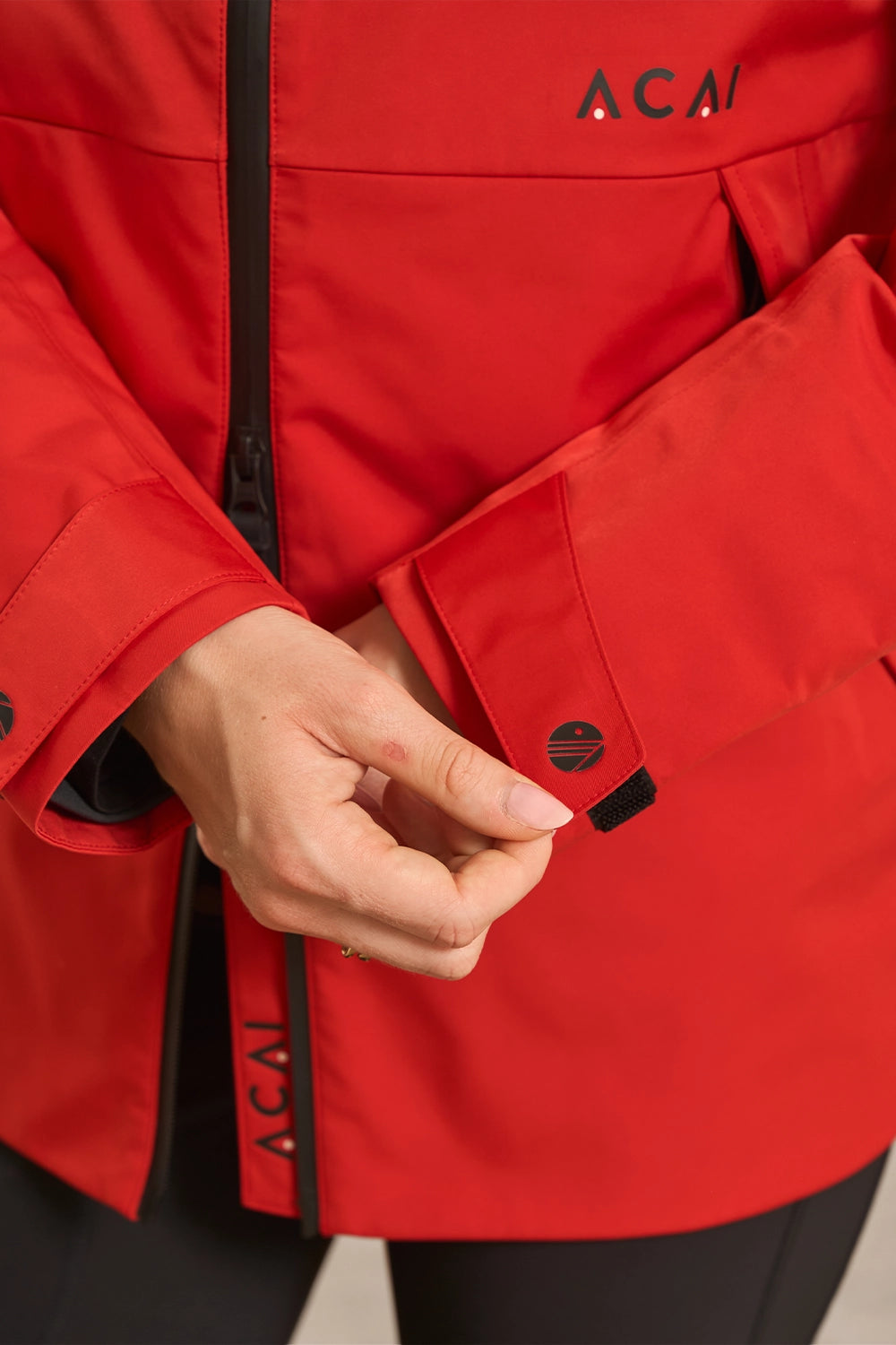 Close-up of a person wearing a bright red Acai jacket, adjusting a flap pocket with a black button tab. The Acai logo is visible on the chest and sleeve.
