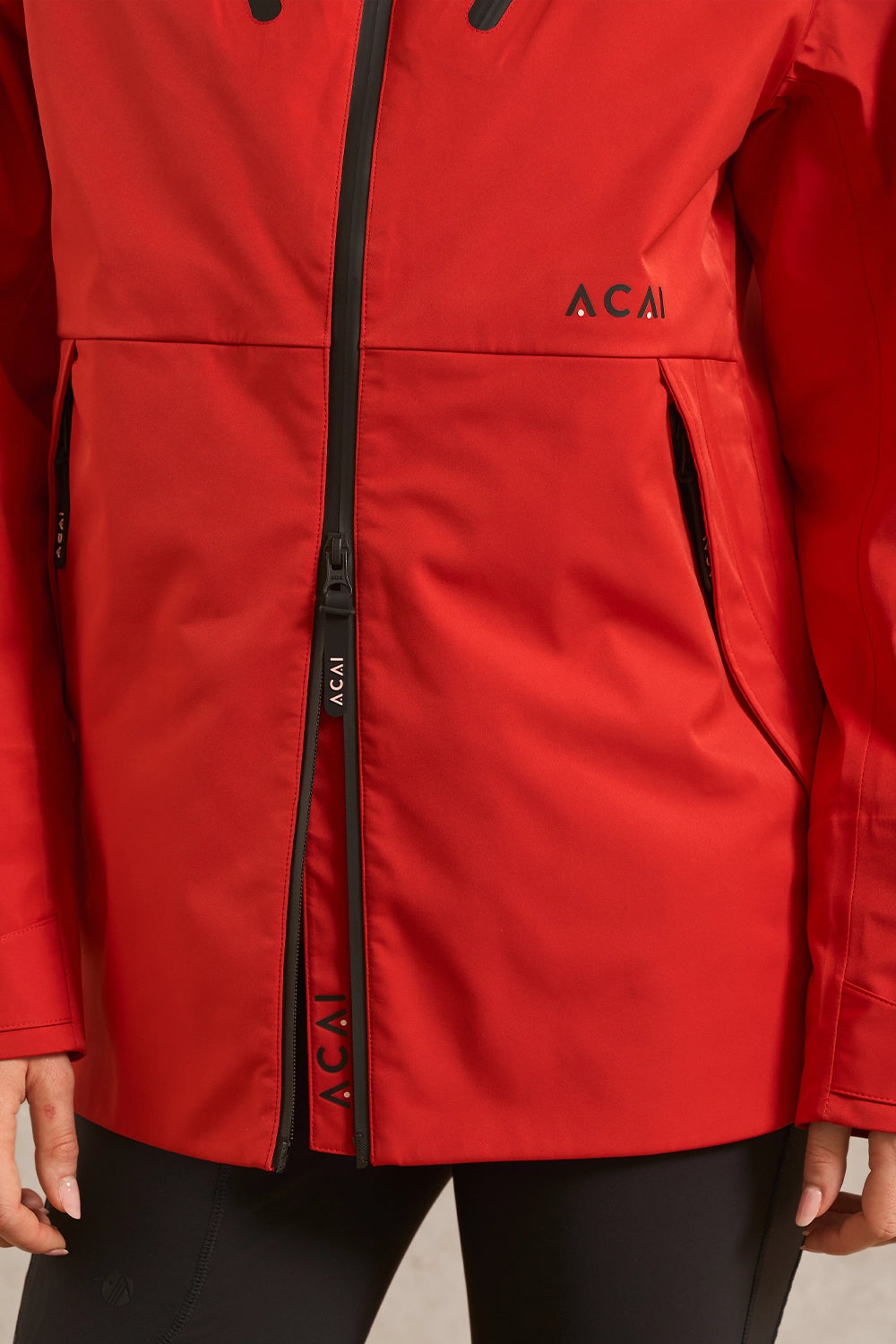 A close-up of a red Acai outdoor jacket with black zippers and black leggings, showing the brand name 'ACAI' printed on the chest and zipper pull.