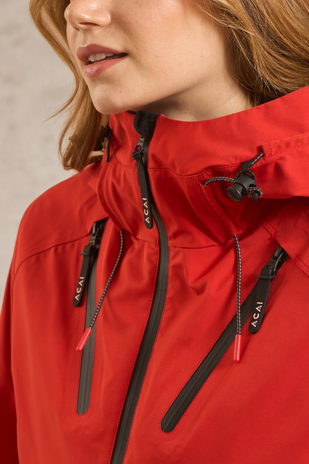 Close-up of a person wearing a bright red technical jacket with black zippers and drawstrings. The zipper pulls are branded with the ACAI logo.