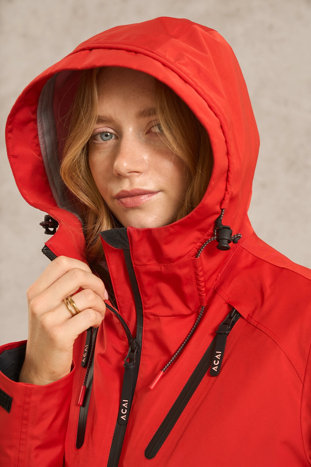 A close-up of a woman wearing a bright red hooded jacket with black zippers and drawstrings, holding the zipper tab. The brand name 'ACAI' is visible on the zipper pulls.