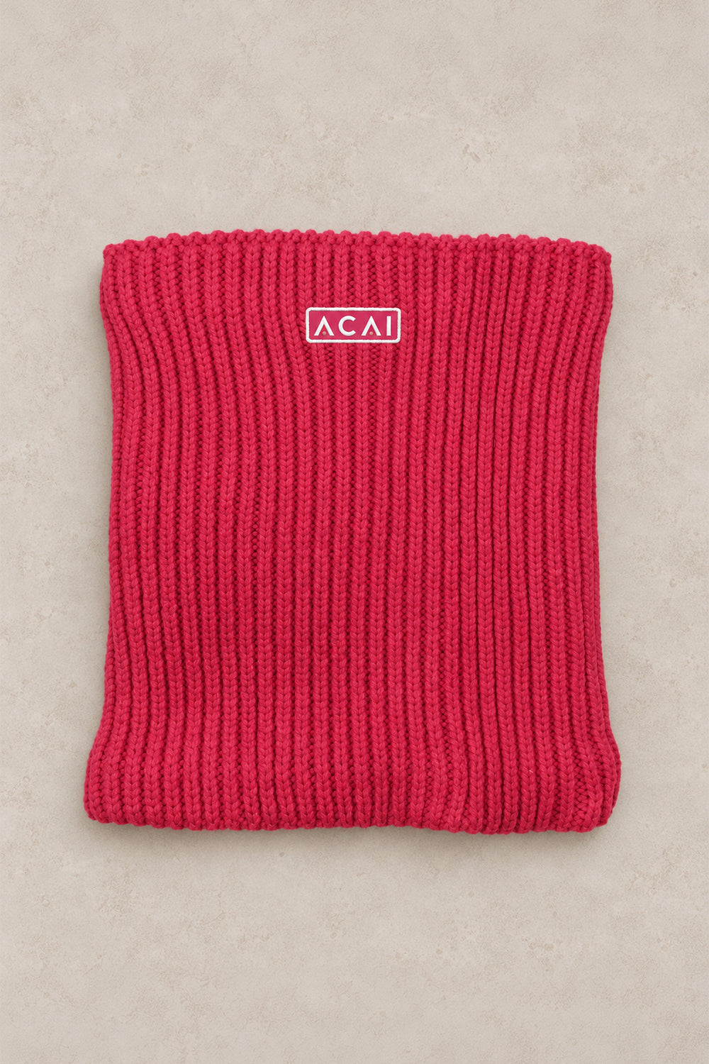 A folded bright pink ribbed knit cowl or neck warmer with a small white rectangular logo patch reading ACAI, placed on a light beige textured background.