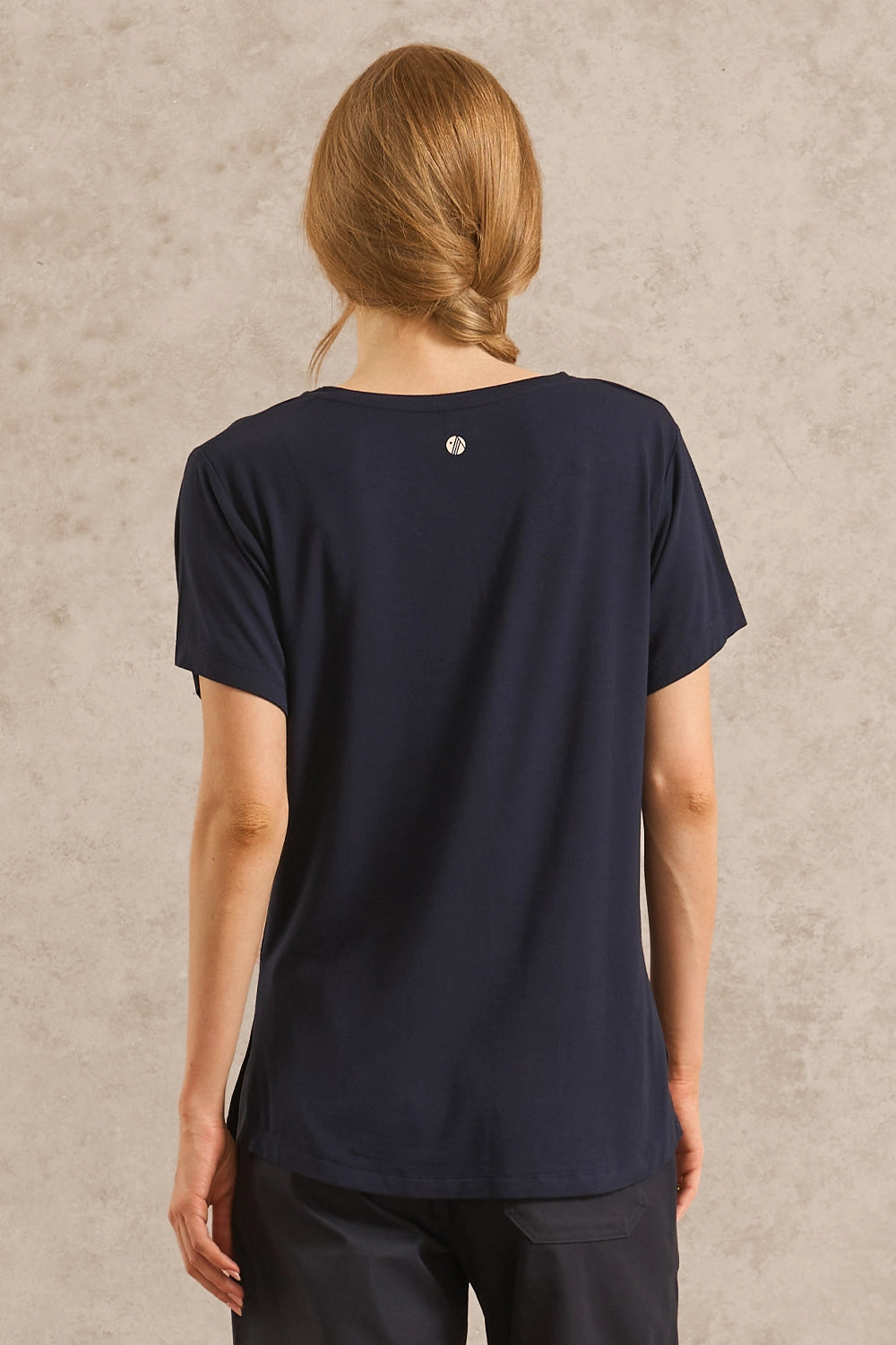 A woman viewed from the back wearing a dark navy blue short-sleeve t-shirt with a small circular silver logo at the center of the back neck, paired with dark pants, set against a textured beige wall.