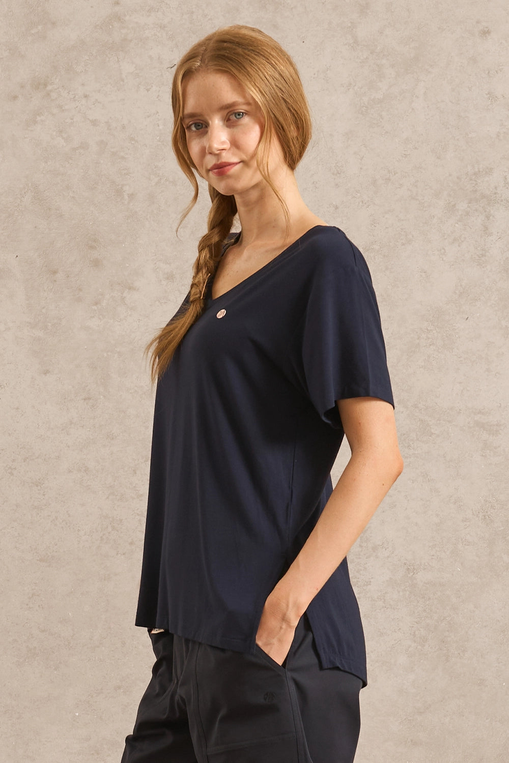 A woman with blonde hair in a braid wearing a navy blue short-sleeve V-neck top and dark pants, standing against a textured beige wall.