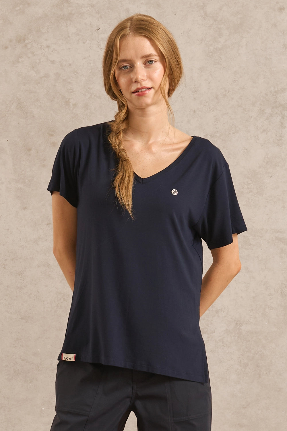 A woman modeling a navy blue short-sleeve V-neck scrub top with a small white circular logo on the left chest, paired with dark pants, against a textured light gray background.