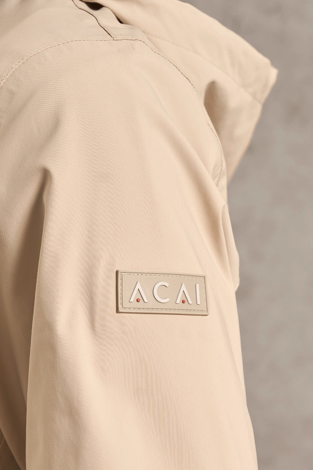 Close-up of a beige jacket sleeve featuring a rectangular patch with the brand name 'ACAI' in white letters and two small red triangles.
