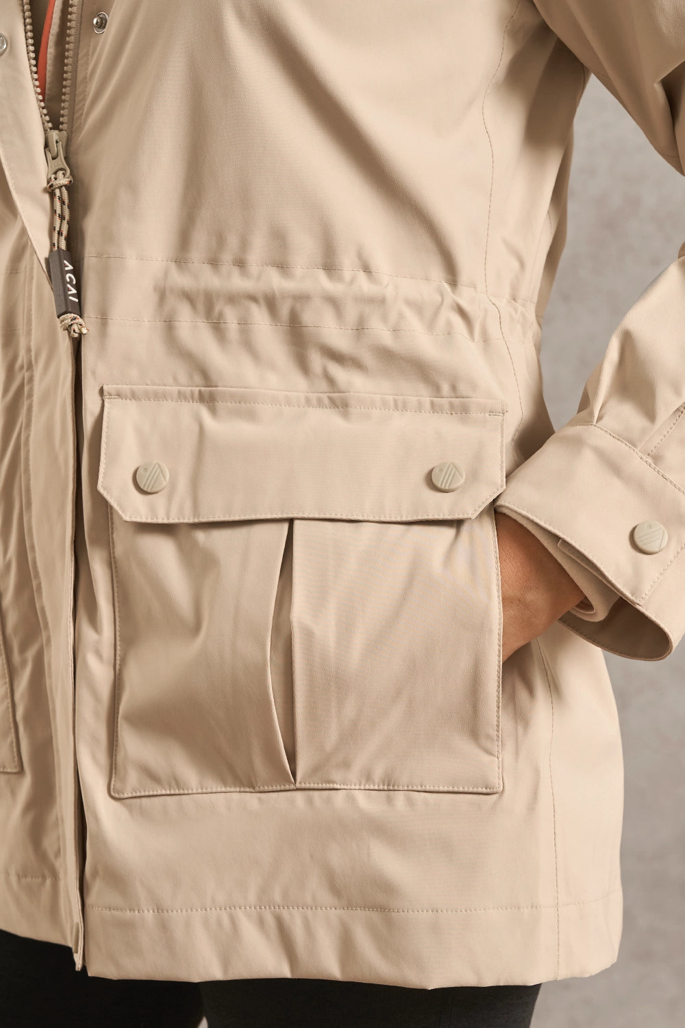 Close-up of a beige jacket with a front flap pocket, snap buttons, and a zipper, worn by a model with one hand in the pocket.