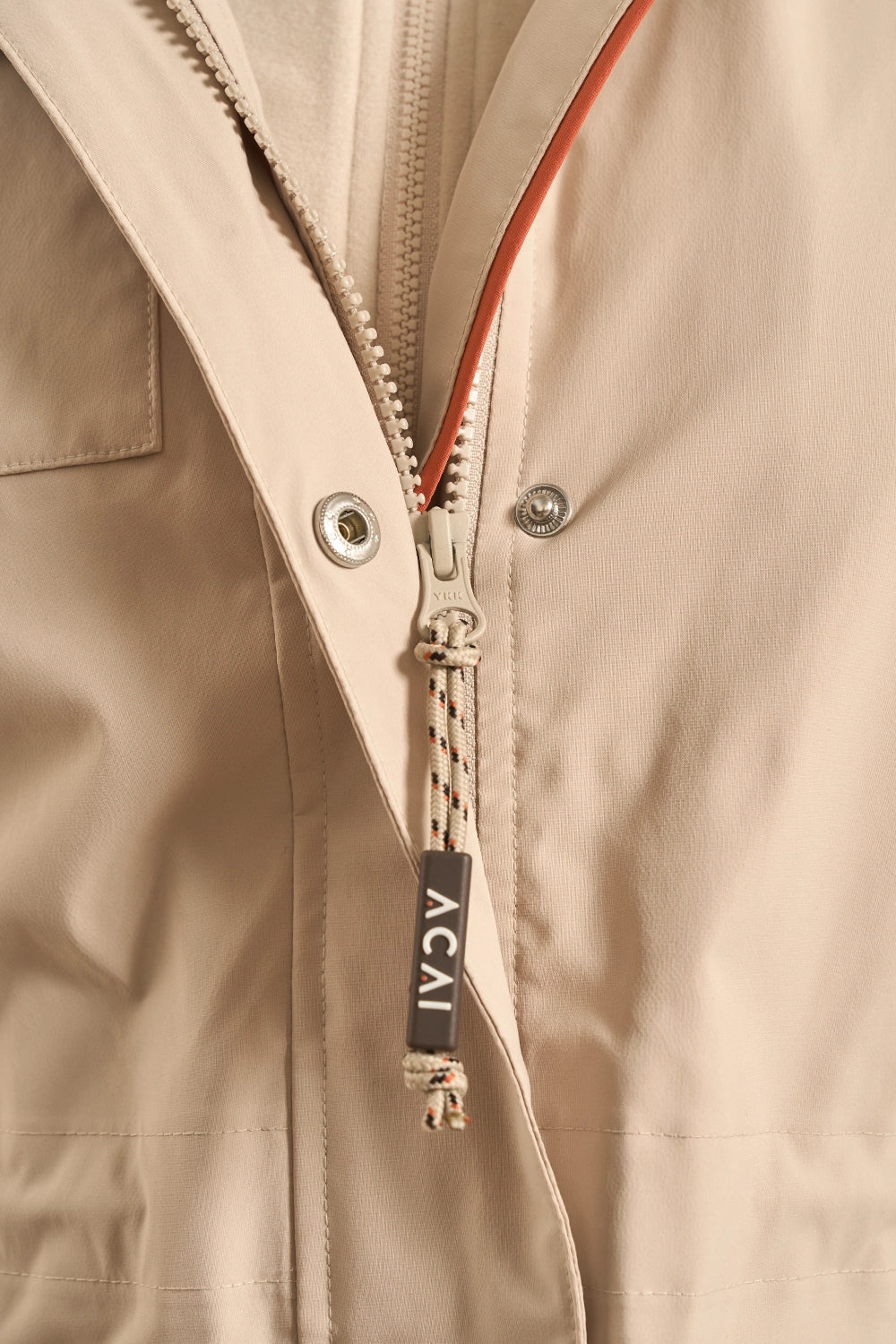 Close-up of a beige zippered jacket with a braided zipper pull featuring a black tag that reads 'ACAI'.