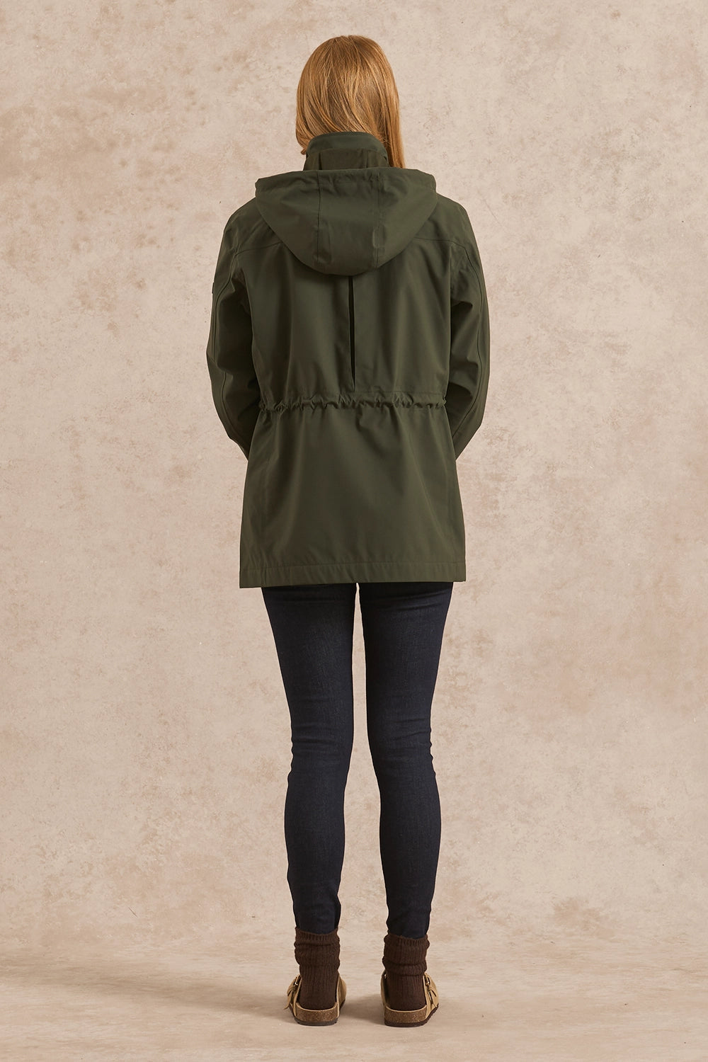 A woman viewed from behind wearing an olive green hooded jacket, dark blue skinny jeans, and brown boots, standing against a textured beige studio backdrop.
