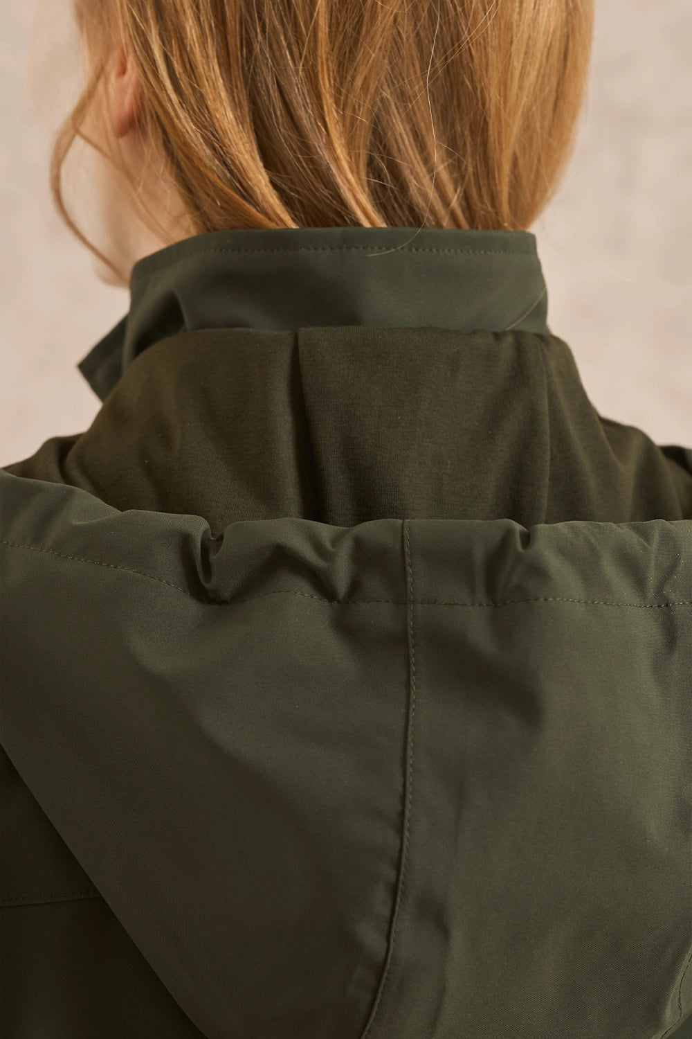 Close-up back view of a person wearing an olive green jacket, showing the collar, shoulder seam, and hair against a soft neutral background.