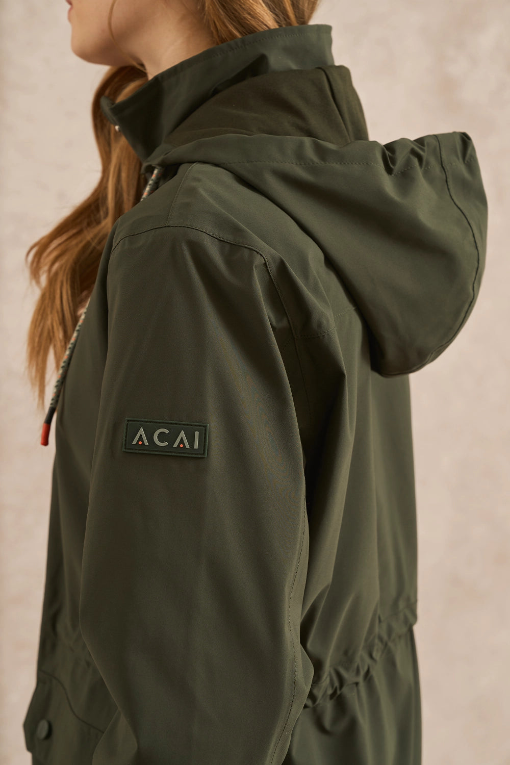 Close-up of a person wearing an olive green hooded jacket, showing the side profile and a black ACAI logo patch on the sleeve, against a neutral beige background.
