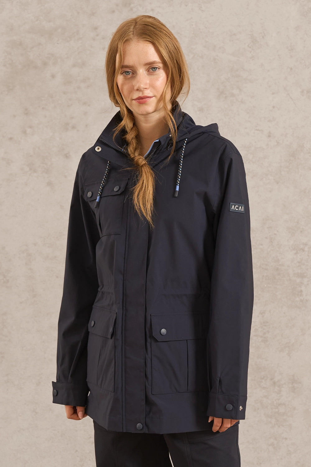 A woman wearing a dark navy hooded jacket with multiple front pockets and drawstrings, standing against a textured light beige wall. The brand name 'ACAI' is visible on the left sleeve.