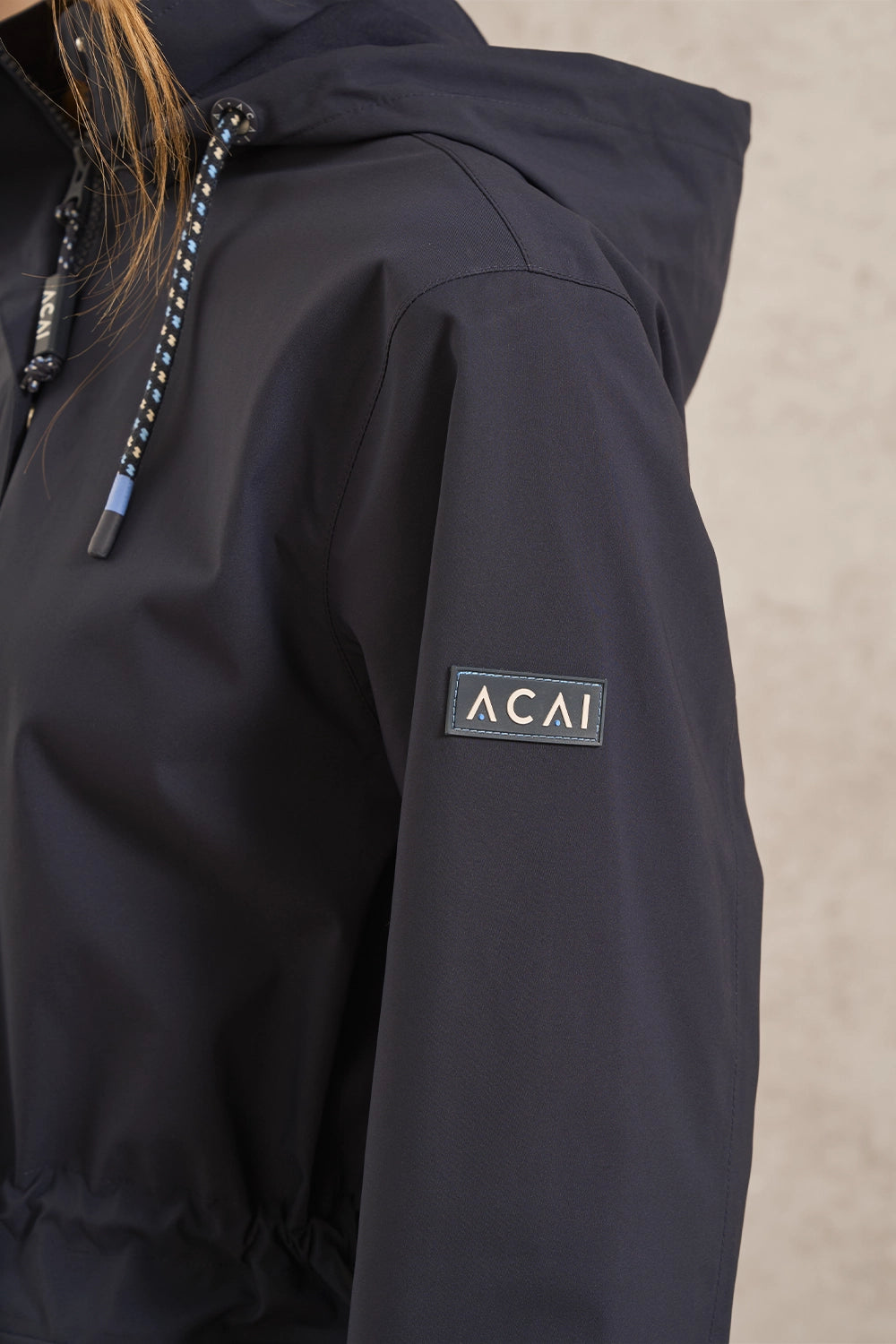 Close-up of a person wearing a dark navy hooded jacket, showing the shoulder, sleeve with a rectangular Acai logo patch, and part of the hood against a light beige background.