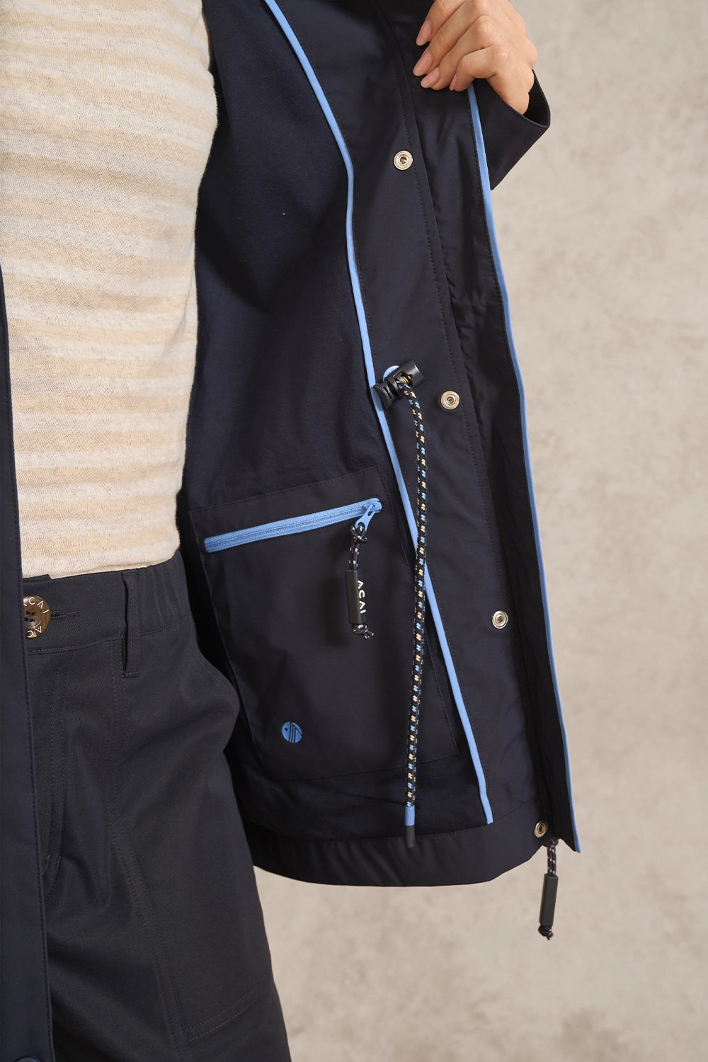 Close-up of a person wearing a dark navy jacket with light blue piping and a matching navy sweater, against a neutral textured background.