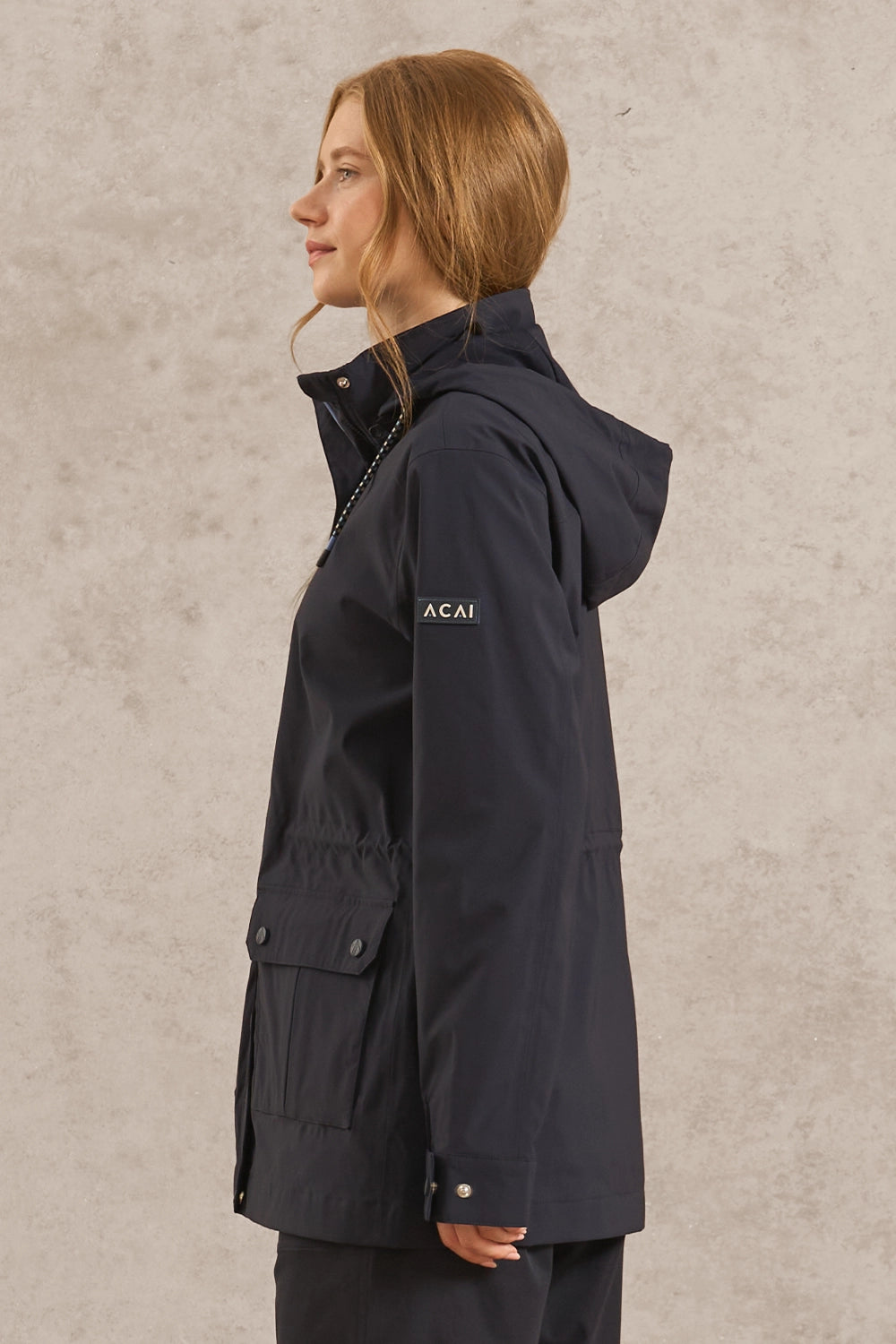 A woman in profile wearing a dark navy hooded jacket with a small white 'ACAI' logo on the chest, set against a textured light gray background.