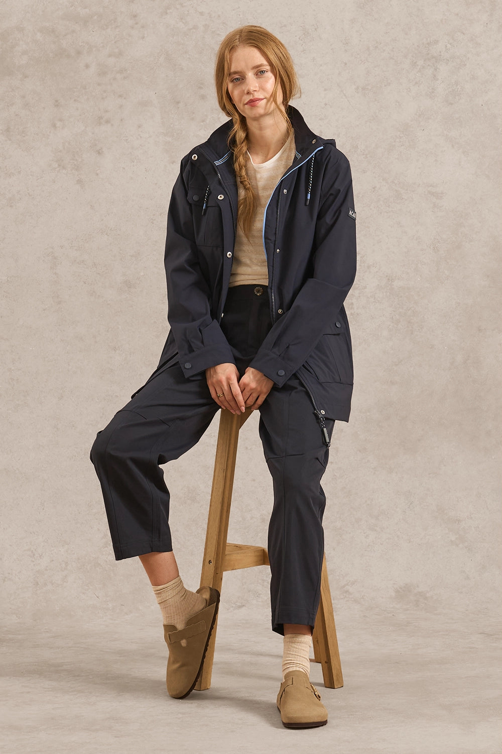 A woman sits on a wooden stool wearing a dark navy hooded jacket, matching navy cropped pants, beige socks, and tan slip-on shoes against a textured light gray studio backdrop.