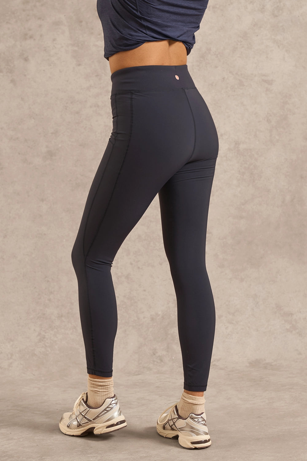 A model wearing high-waisted dark navy athletic leggings, paired with beige socks and white sneakers, shown from a side-rear angle against a textured gray studio backdrop.