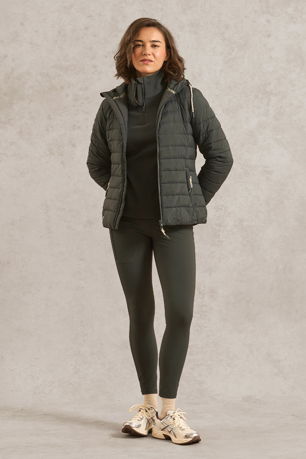 A woman modeling a dark gray quilted puffer jacket over a matching dark gray turtleneck and leggings, paired with beige sneakers, against a textured gray studio backdrop.