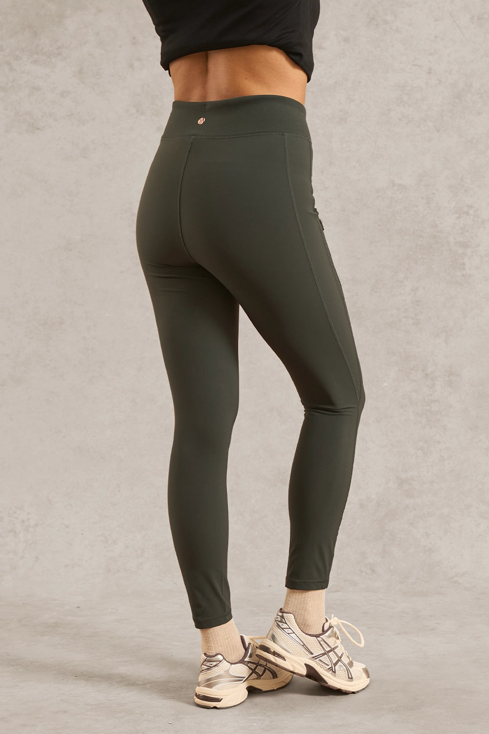 A person wearing dark olive green high-waisted leggings, shown from the back, standing against a textured gray studio wall. The person is also wearing a black cropped top and beige athletic sneakers.