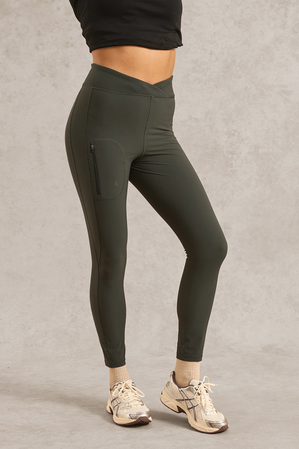 A person modeling dark olive green high-waisted leggings with a zippered side pocket, paired with beige sneakers and a black cropped top, standing against a textured gray studio background.
