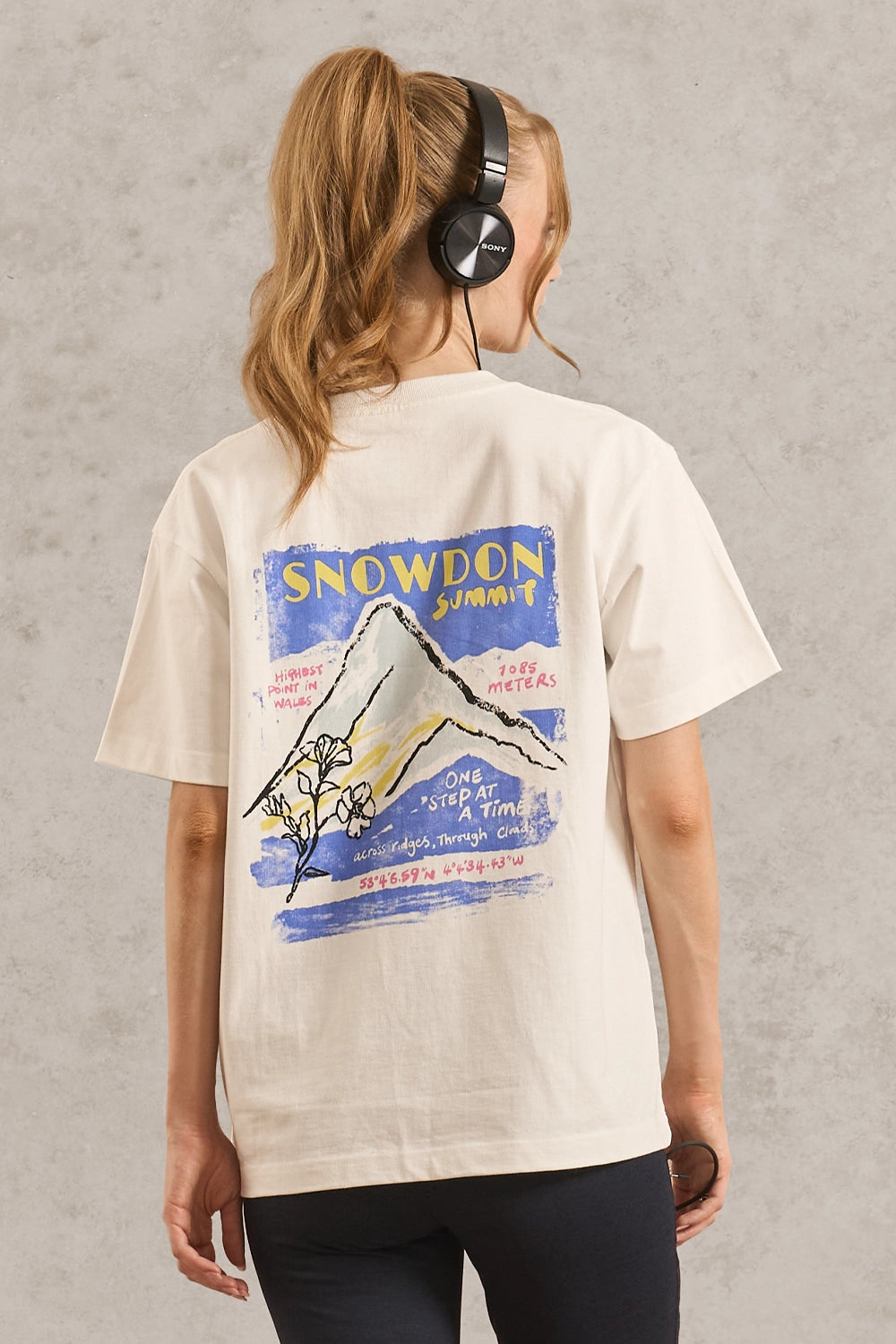 A woman viewed from behind wearing a cream-colored t-shirt with a large graphic print of a mountain and the text 'SNOWDON SUMMIT' on the back, paired with black pants against a textured gray wall.