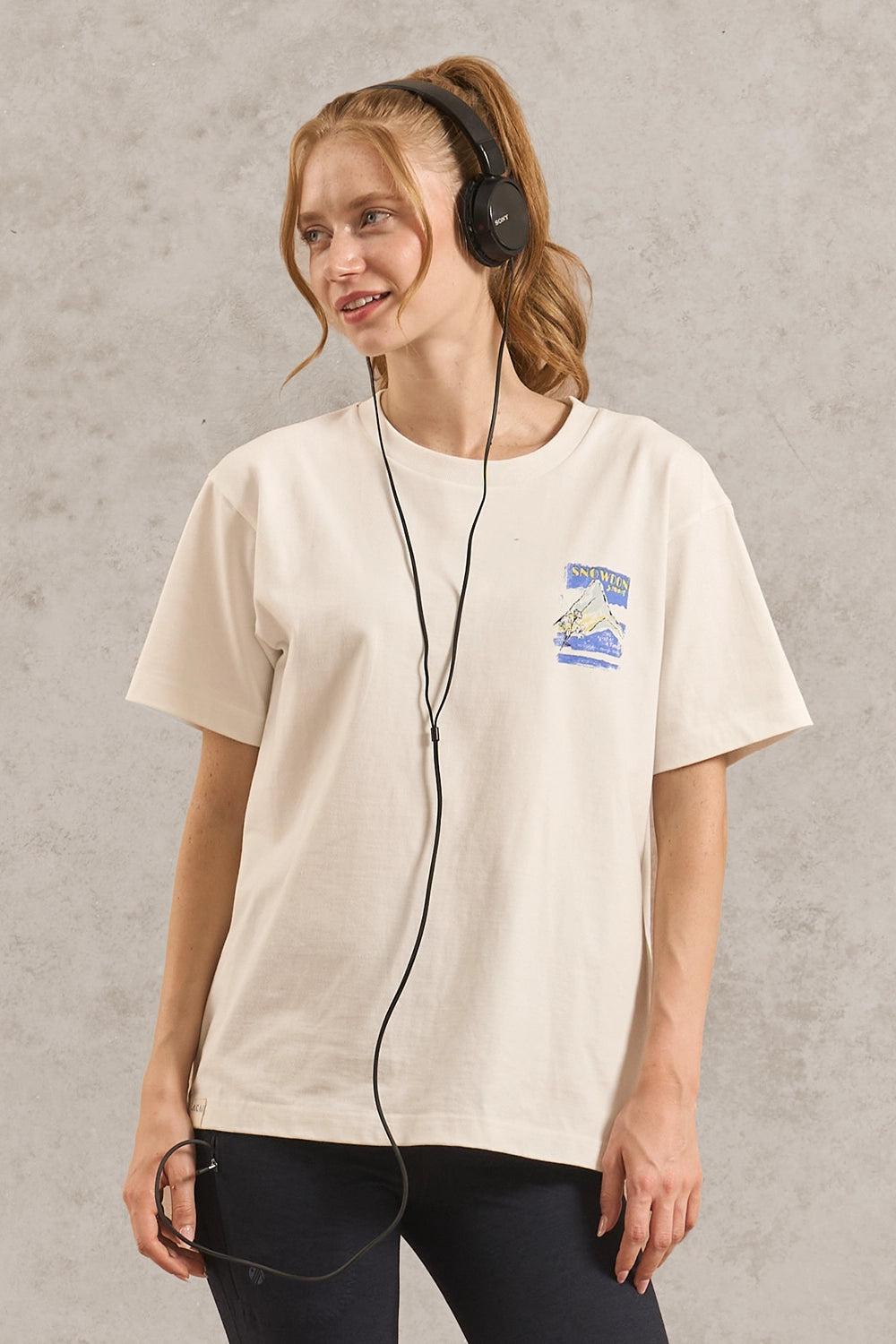 A woman wearing a cream-colored short-sleeve t-shirt with a small blue and white graphic on the left chest, paired with black leggings and black over-ear headphones, standing against a textured gray wall.
