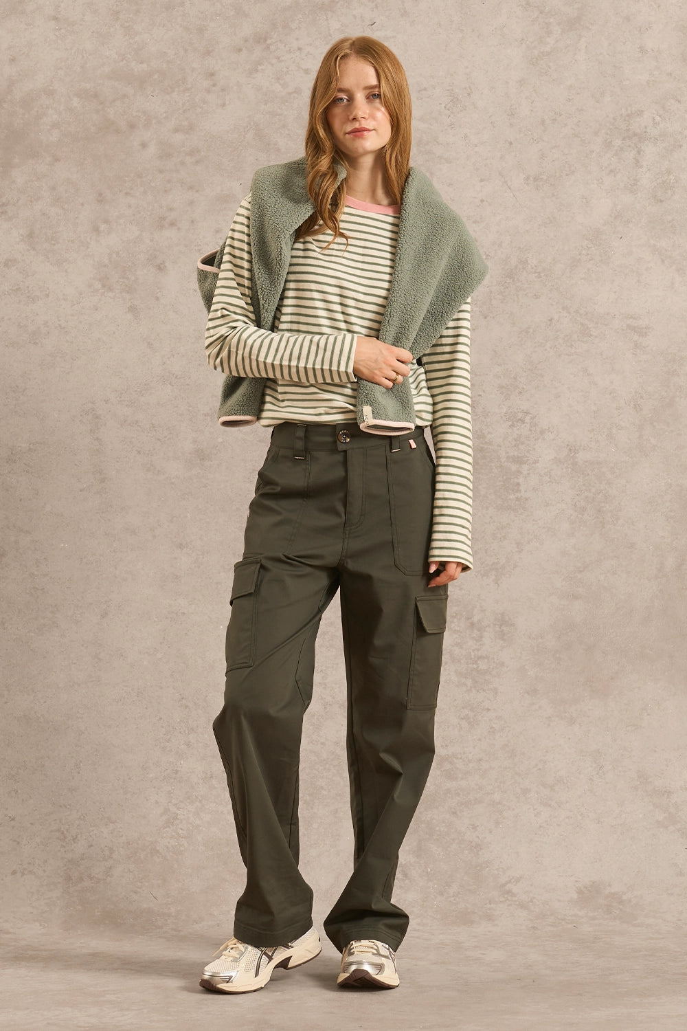 A model stands against a textured beige studio backdrop wearing a striped long-sleeve top, olive green cargo pants, a light green knit cardigan draped over her shoulders, and white sneakers.