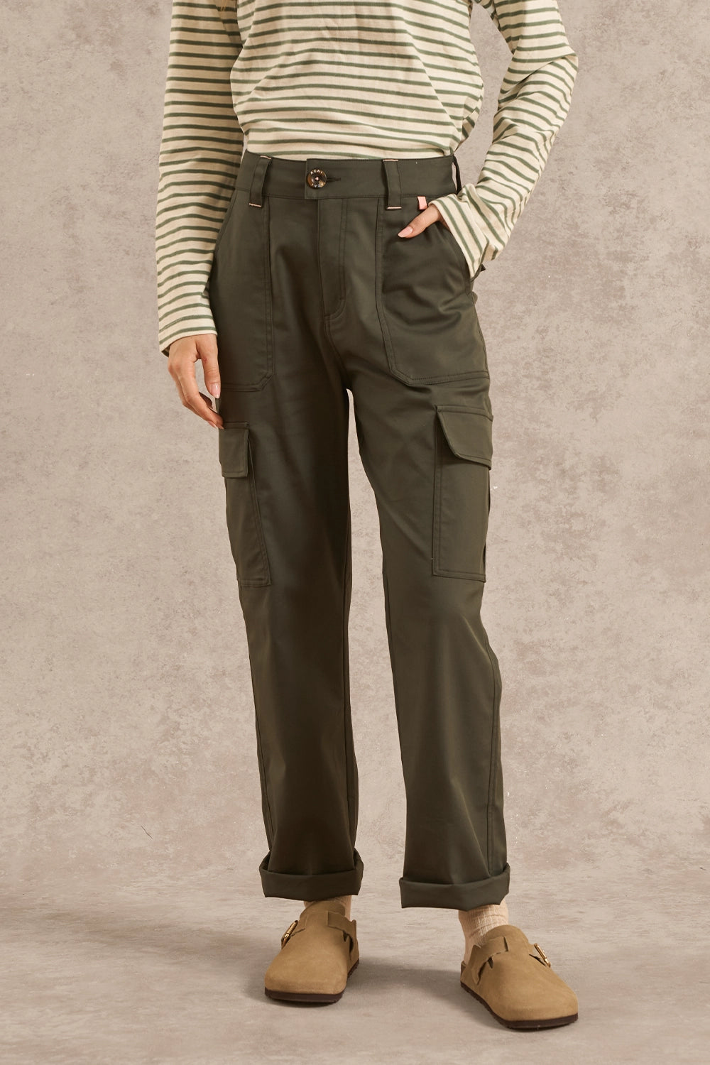 A person wearing olive green cargo pants with a striped long-sleeve shirt and tan slip-on shoes, standing against a textured beige studio backdrop.
