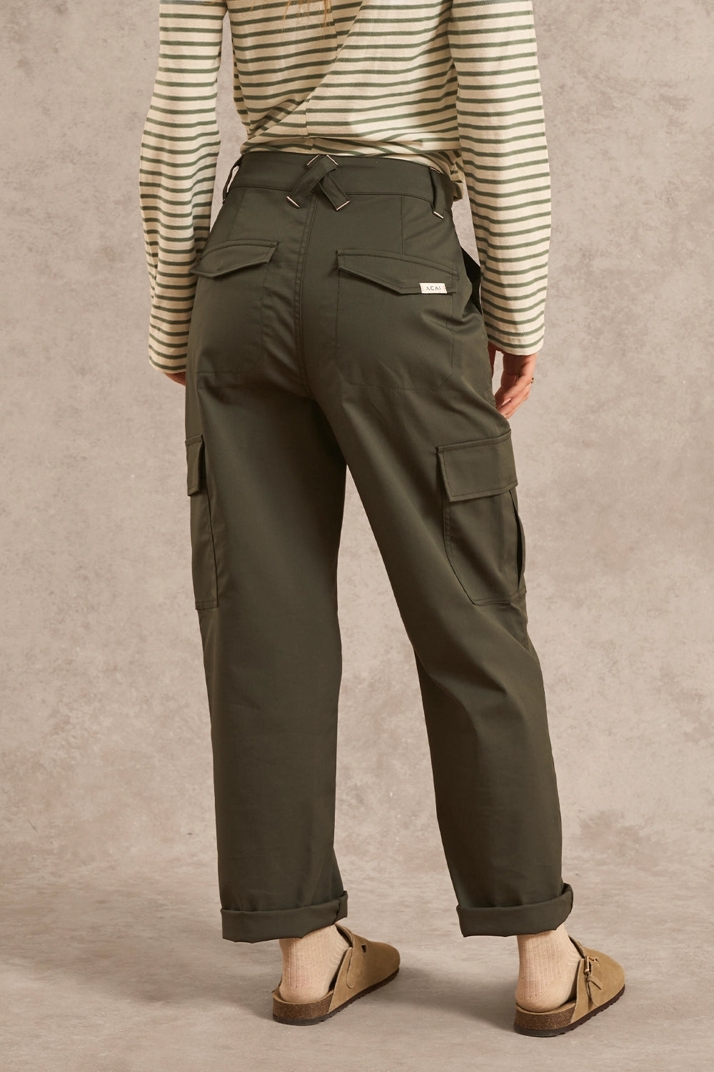 A person shown from the back wearing olive green cargo pants, a cream and green striped long-sleeve top, and tan mule sandals against a textured beige studio backdrop.