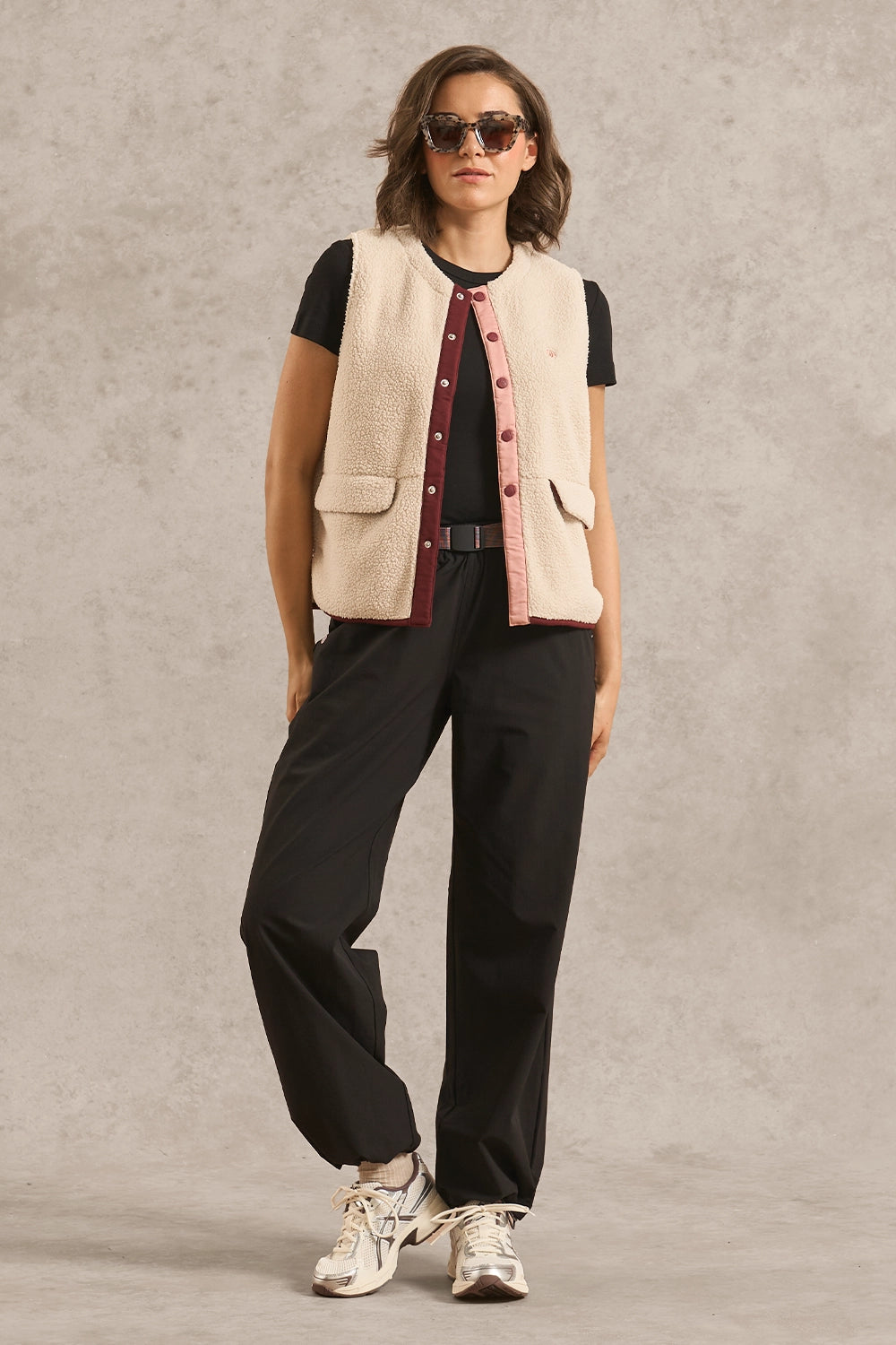 A model wearing a cream-colored textured vest over a black t-shirt with a burgundy striped tie detail, paired with black trousers and white sneakers, set against a textured gray studio background.