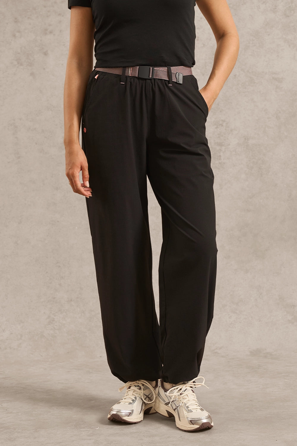 A person wearing black wide-leg jogger-style pants with a drawstring waist, paired with a dark belt, black top, and white sneakers, standing against a textured gray studio backdrop.
