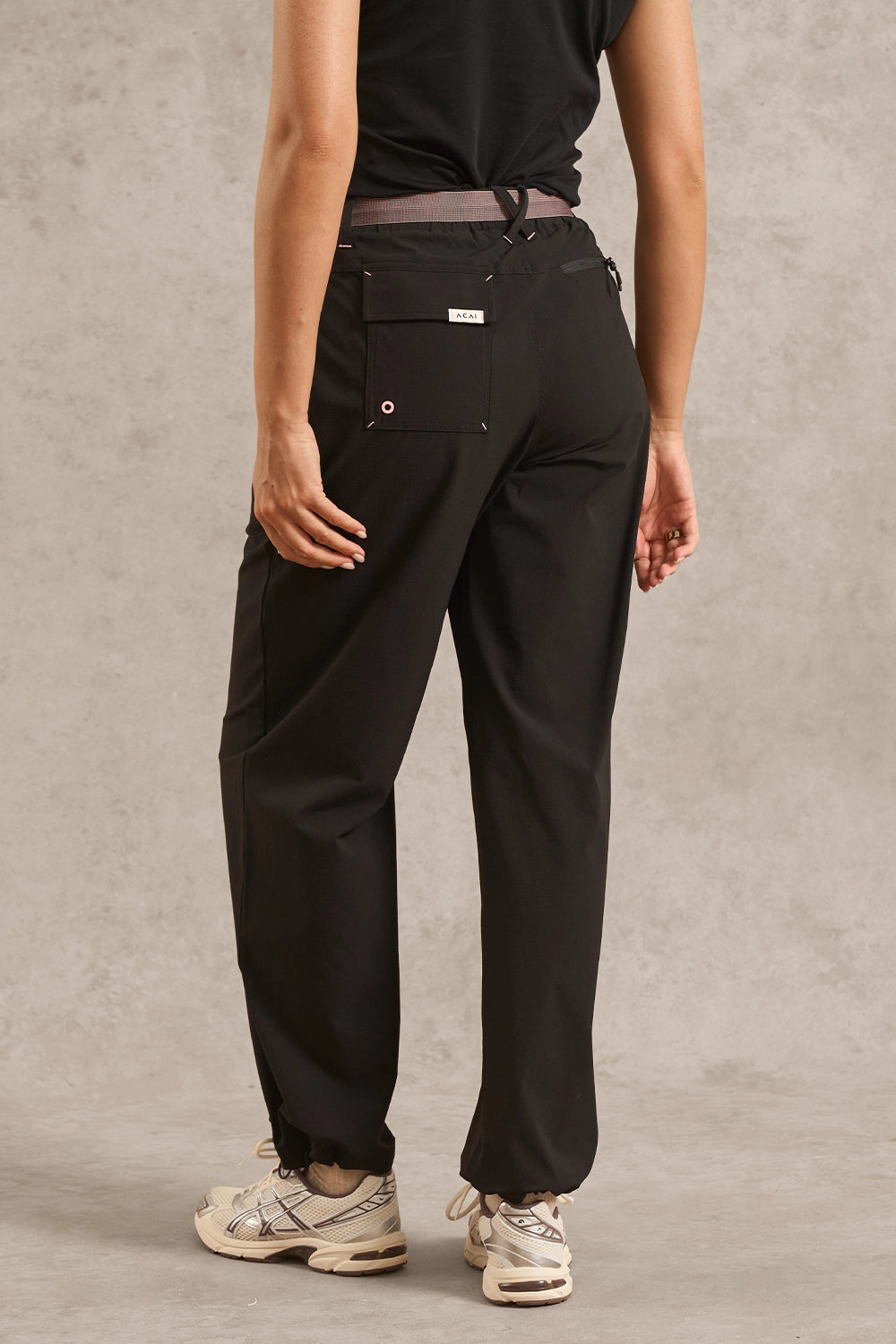 A person shown from the back wearing loose black pants with a drawstring waist, a black top, and white sneakers against a textured gray studio backdrop.