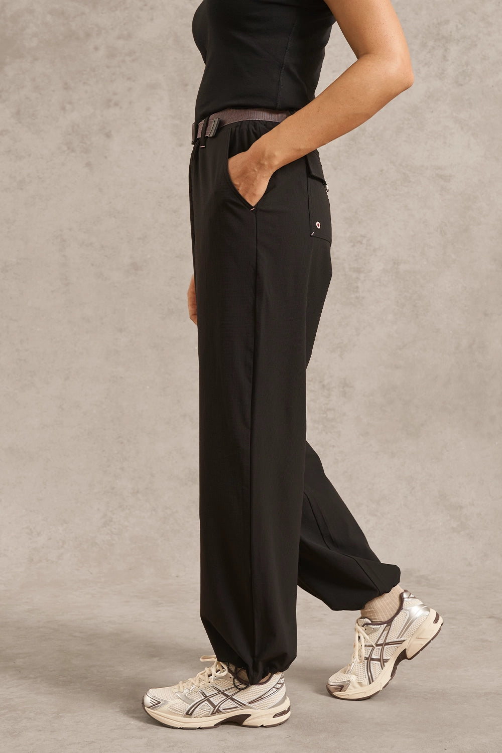 A person wearing loose black wide-leg pants with a dark belt, black top, and white athletic sneakers, standing against a textured gray studio backdrop.