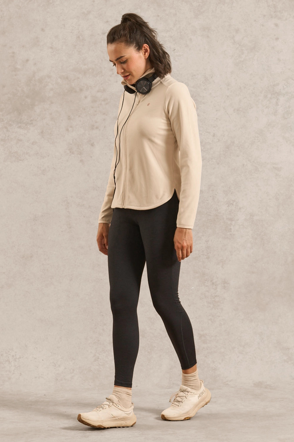 A woman modeling a light beige long-sleeve athletic zip-up jacket and black leggings, standing against a textured light gray studio backdrop.