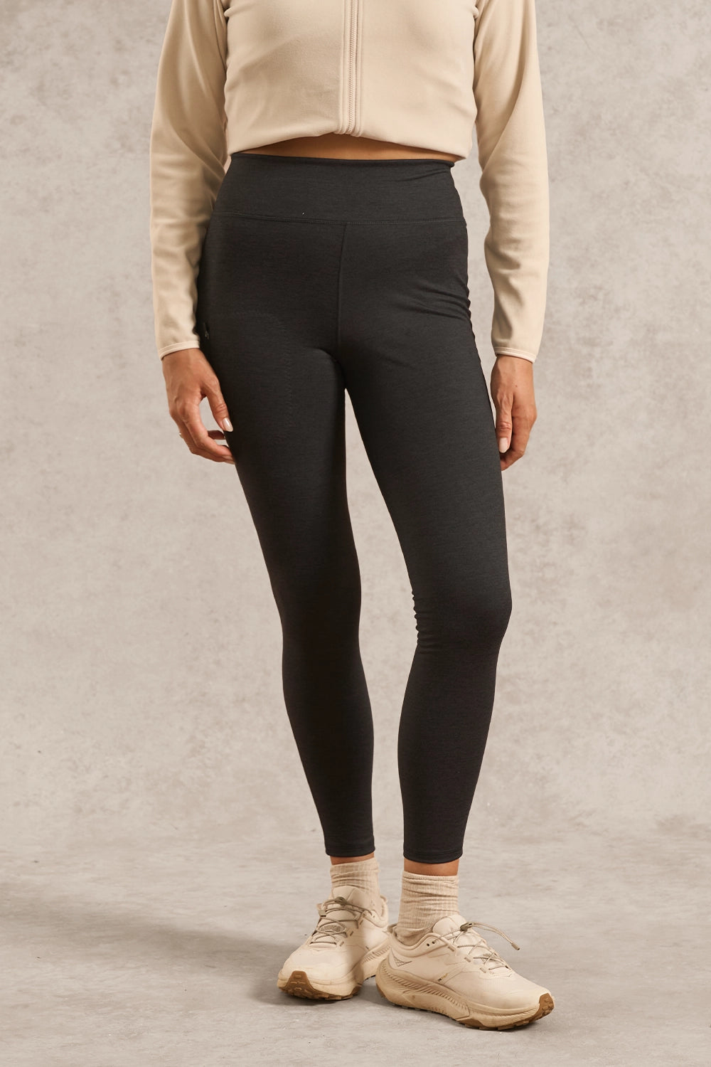 A model wearing black high-waisted athletic leggings, a beige cropped zip-up jacket, beige socks, and beige sneakers, standing against a textured gray studio backdrop.