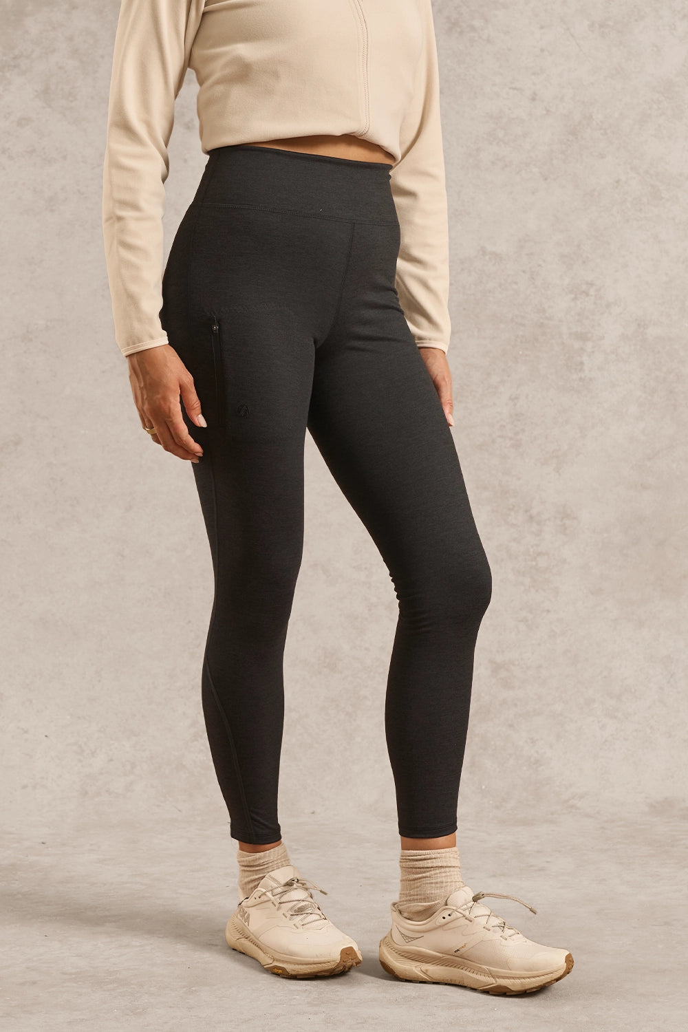 A model wearing high-waisted black leggings, a beige cropped sweatshirt, beige socks, and beige sneakers, standing against a textured gray studio background.