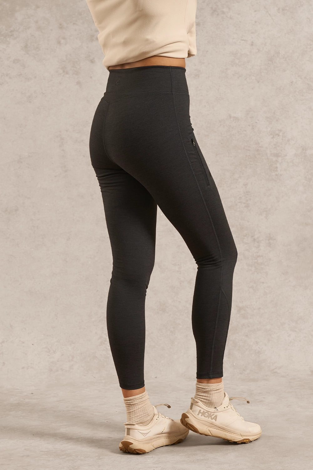 A person wearing black high-waisted leggings, paired with a beige sweatshirt, beige socks, and beige sneakers, standing against a textured gray studio backdrop.