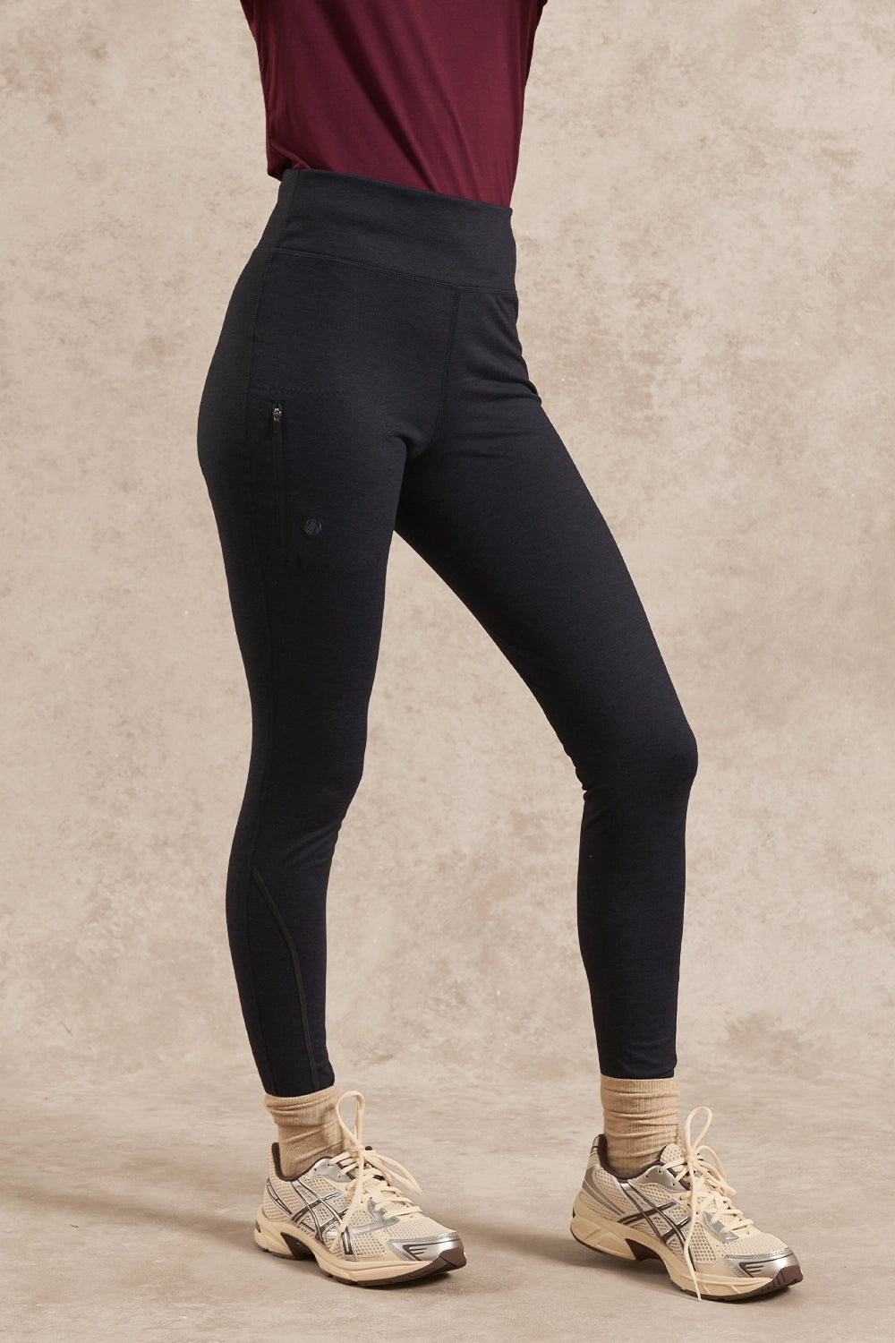 A person wearing high-waisted black athletic leggings with a zippered pocket, paired with beige socks and running shoes, standing against a textured beige studio backdrop.
