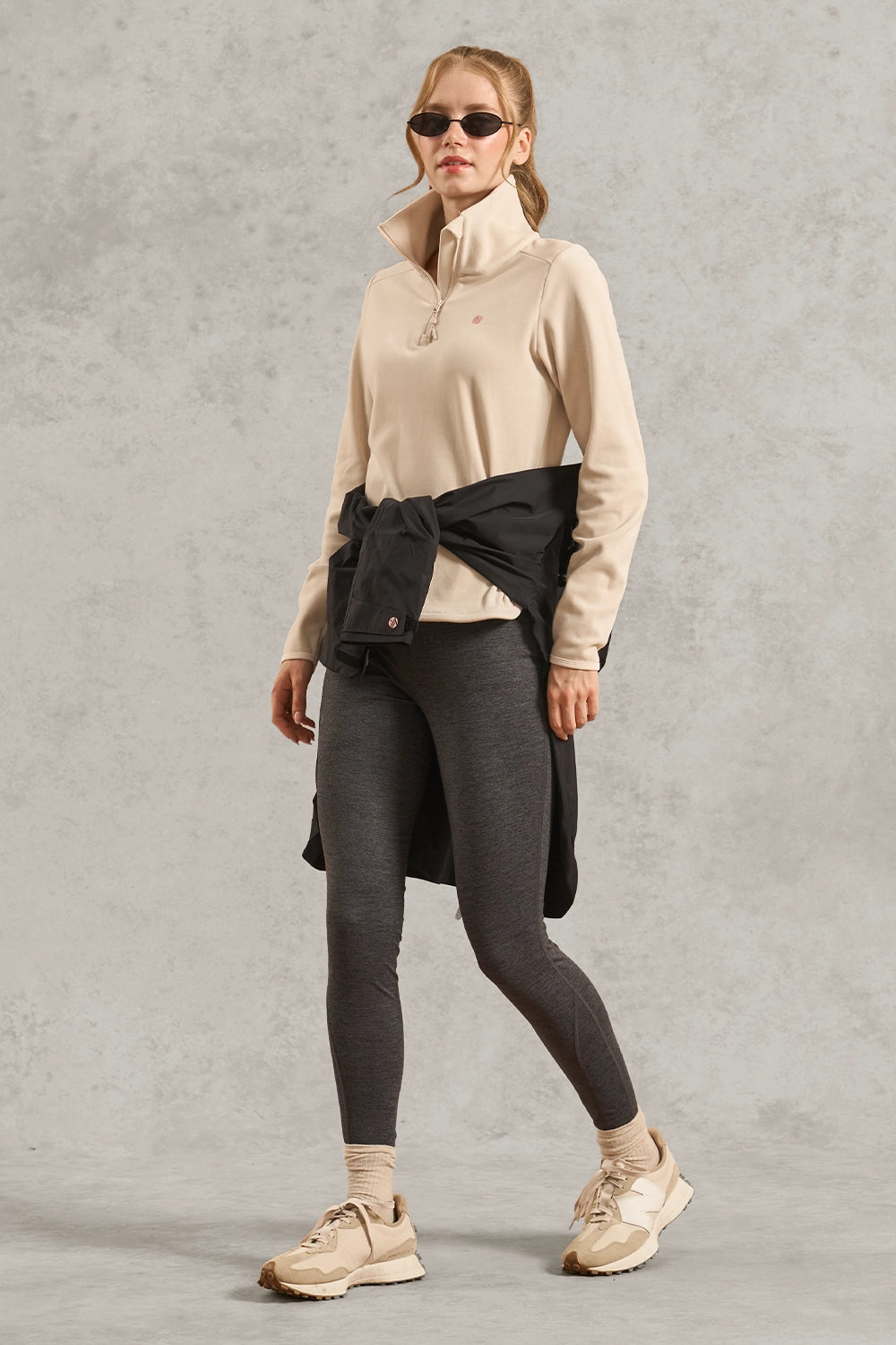 A woman modeling a beige quarter-zip pullover, dark gray leggings, and beige sneakers, with a black jacket tied around her waist, set against a textured gray studio background.